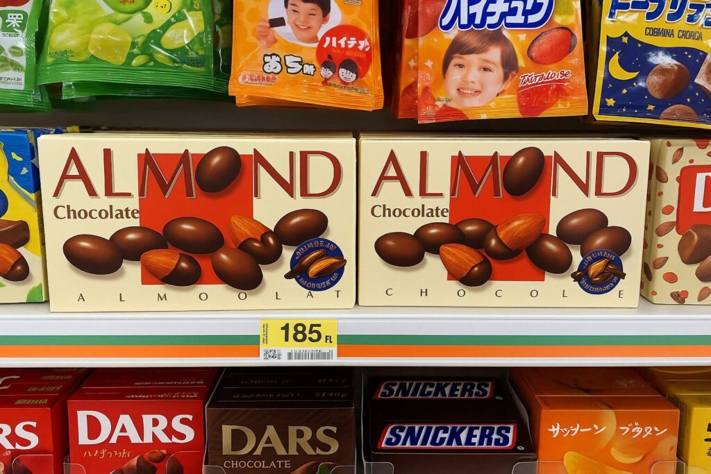 Meiji Almond Chocolate boxes displayed on a Japanese convenience store shelf alongside other popular snacks like DARS, Hi-Chew, and Snickers.